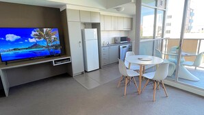 A modern kitchen with white cabinets, a refrigerator, and a microwave. There is a dining area with a round table and white chairs. A flat-screen TV is mounted on the wall, displaying a tropical scene.