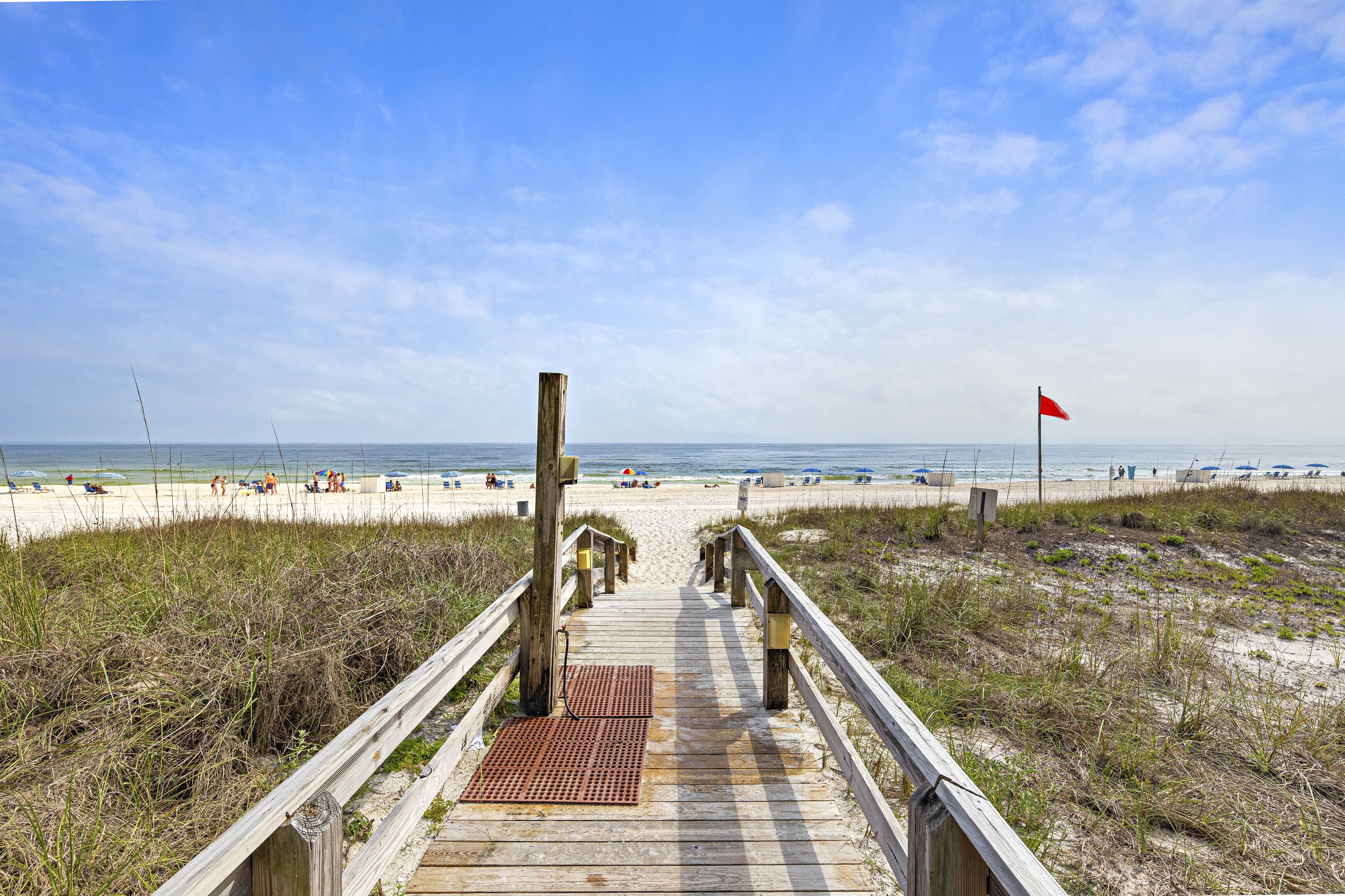 A wooden boardwalk leading to a beach with a red flag, sandy dunes, and clear blue sky.
