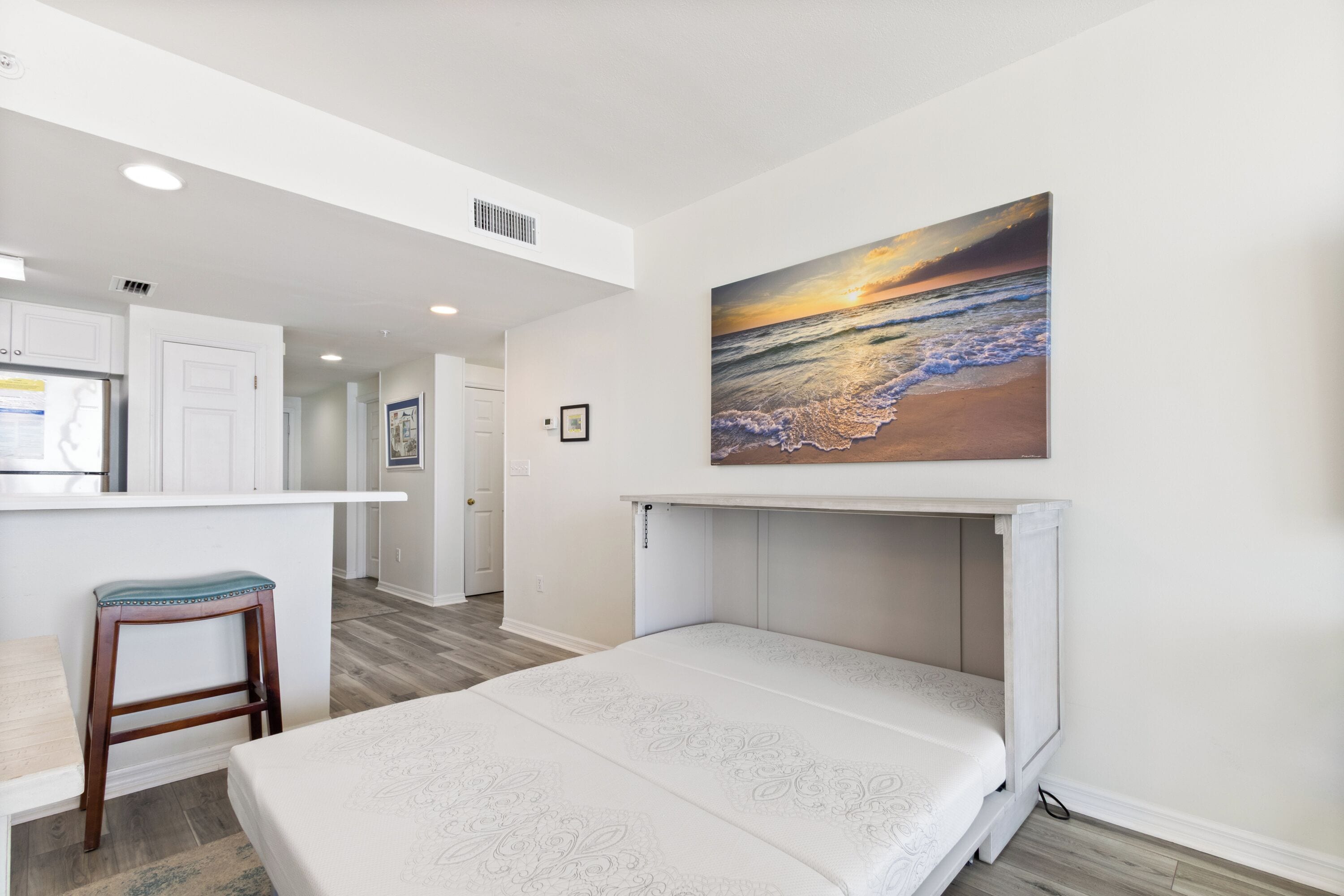A bedroom with a bed, a painting of a beach scene, a kitchenette, and a bathroom.