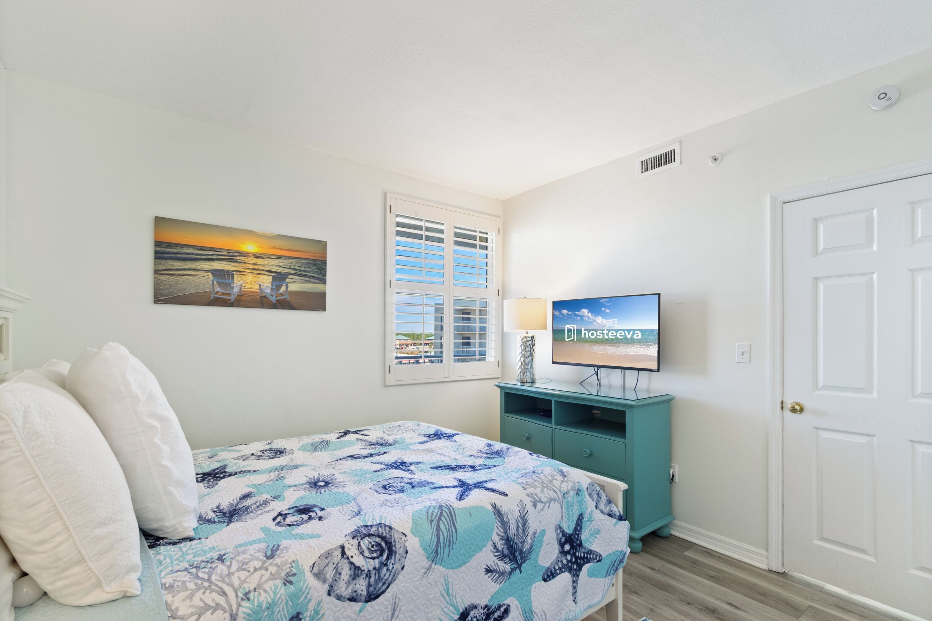 A bedroom with a bed, a television, a window with shutters, and a painting of a sunset.