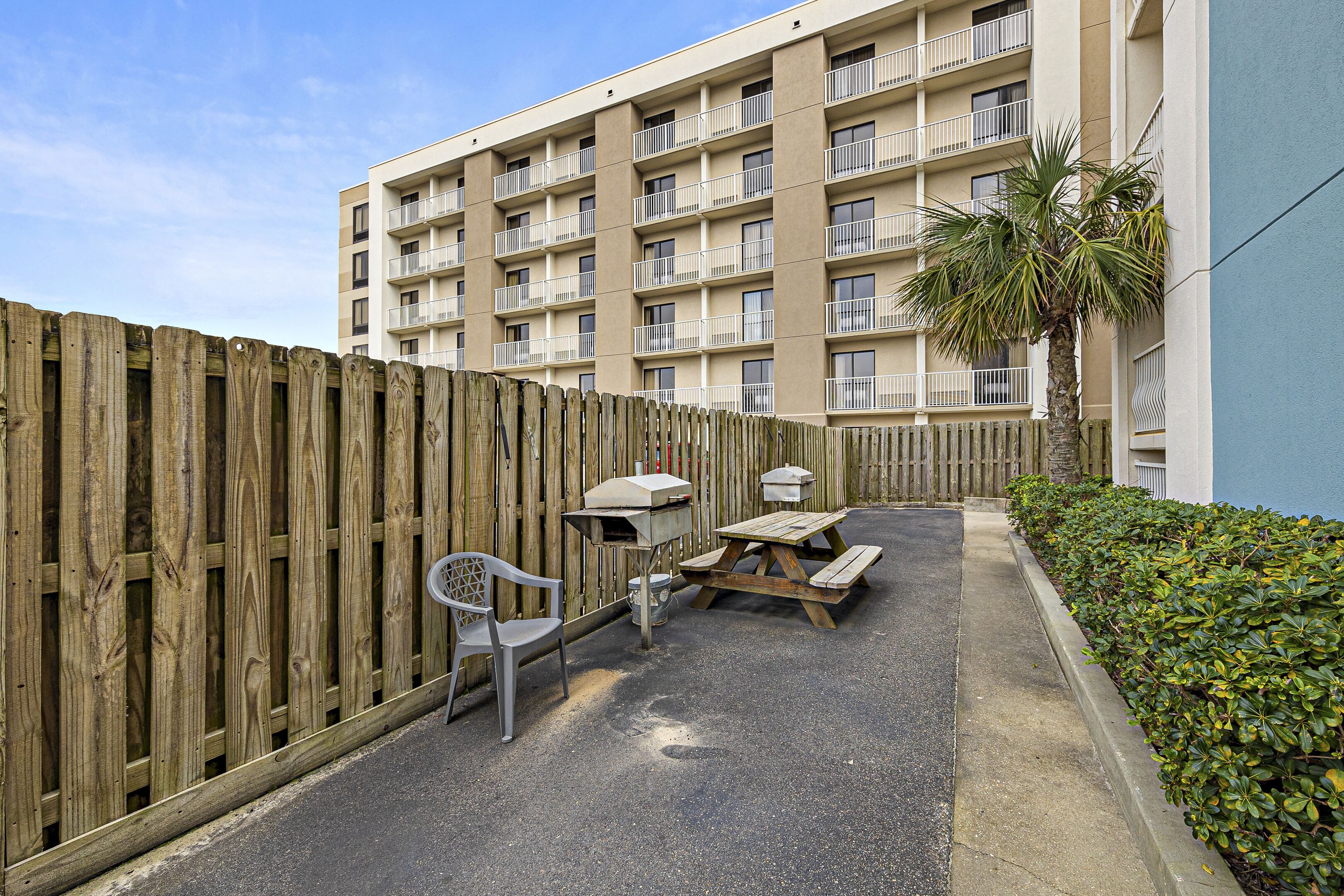 A wooden fence, a picnic table, a grill, and a chair in an outdoor area with a building in the background.
