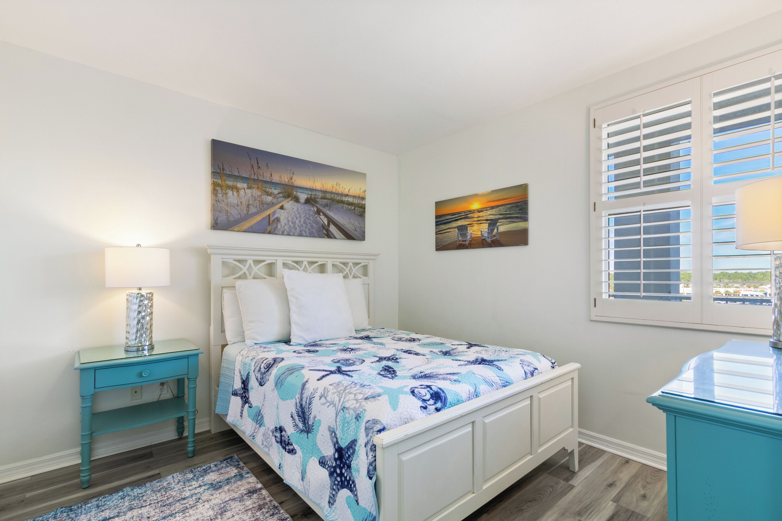 A bedroom with a bed, a nightstand, a lamp, a window with blinds, and two wall-mounted paintings.