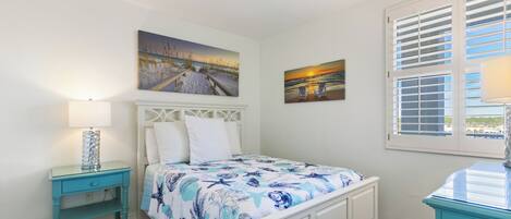 A bedroom with a bed, a nightstand, a lamp, a window with blinds, and two wall-mounted paintings.