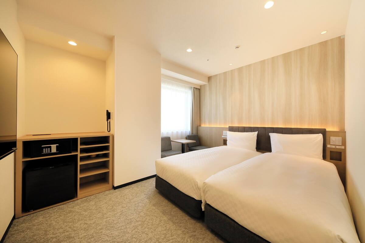 A hotel room with two beds, a desk, a chair, a small table, and a built-in wardrobe.