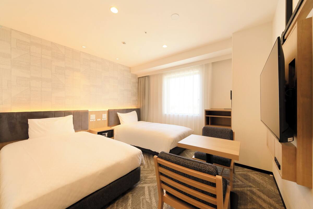 A hotel room with two beds, a desk, a chair, a TV, and a large window.
