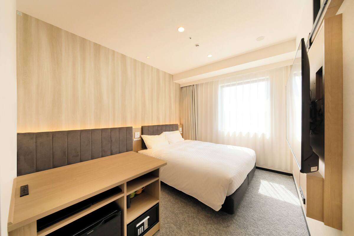 A hotel room with a bed, a TV mounted on the wall, a wooden desk, and a window with curtains.