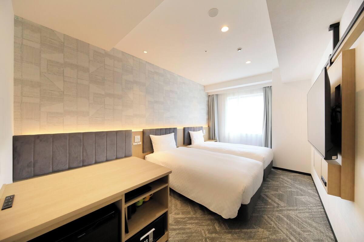 A hotel room with two beds, a desk, a TV, and a large window.