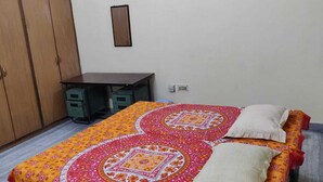 A bedroom with a bed, a wooden wardrobe, a desk, and a small cabinet. - A perfect stay for family & friends. (VARANASI)