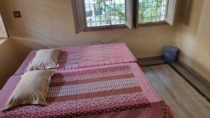 A small, simple room with two beds, a window with bars, a blue bucket, and a plain wall. - A perfect stay for family & friends. (VARANASI)