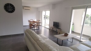 En moderne stue med en hvid sofa, et spisebord med stole, et fladskærms-tv på et stativ, og et stort vindue der giver naturligt lys.