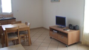 A small, clean room with a wooden dining table, chairs, a TV on a wooden stand, a small cabinet, and a window with curtains. - Beautiful independent cottage with garden (Contes)