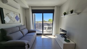 Ein modernes Wohnzimmer mit grauem Sofa, einem Flachbildfernseher auf einem Schrank und einem Balkon mit Tisch und Stühlen.