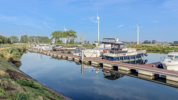 Una vía fluvial tranquila con barcos amarrados a la orilla, rodeada de vegetación y turbinas eólicas al fondo.