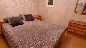 Siististi pedattu sänky, jossa on kukkakuviollinen päiväpeite, kaksi tyynyä, yöpöytälamppu ja seinällä oleva kehystetty kuva.