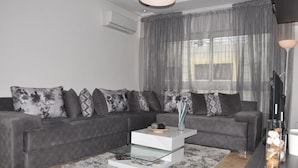 Ein modernes Wohnzimmer mit grauem Sofa, einem Glastisch und einem großen Fenster mit durchsichtigen Vorhängen.