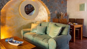 A cozy living room with a green sofa, a wooden coffee table, and a stone wall with a circular window. - Carob Cottage (Plaka Elounda)