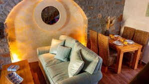 A cozy living area with a stone wall, a round mirror, a sofa with cushions, a wooden coffee table, and a dining table set for two. - Carob Cottage (Plaka Elounda)