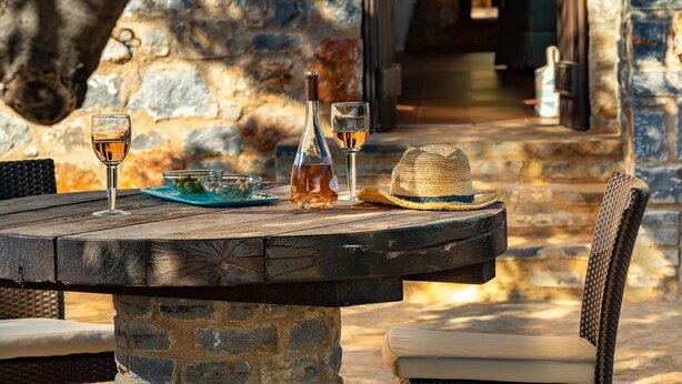 A stone table with a bottle and glasses, a wicker chair, and a stone wall in the background. - Carob Cottage (Plaka Elounda)