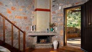 A stone fireplace with a wooden staircase, a wooden table with a bowl, and a door leading to a garden. - Carob Cottage (Plaka Elounda)