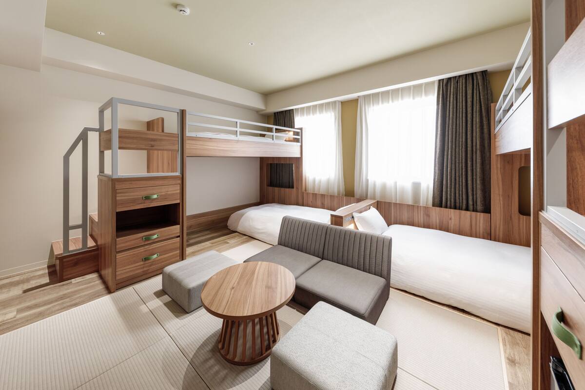 Ein modernes Hotelzimmer mit Etagenbett, Sofa, Hocker und einem kleinen Holztisch.