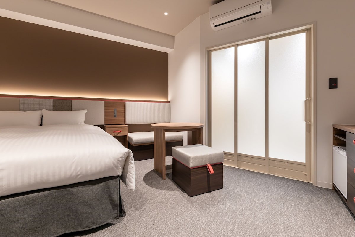 Ein modernes Hotelzimmer mit einem großen Bett, einem kleinen Tisch, einer Bank, einem Kleiderschrank und einer Klimaanlage.