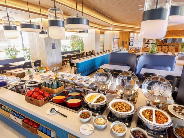 Ein Buffet mit verschiedenen Speisen und Besteck, eine Sitzecke mit Tischen und Stühlen sowie eine Theke mit blauer Wandverkleidung.