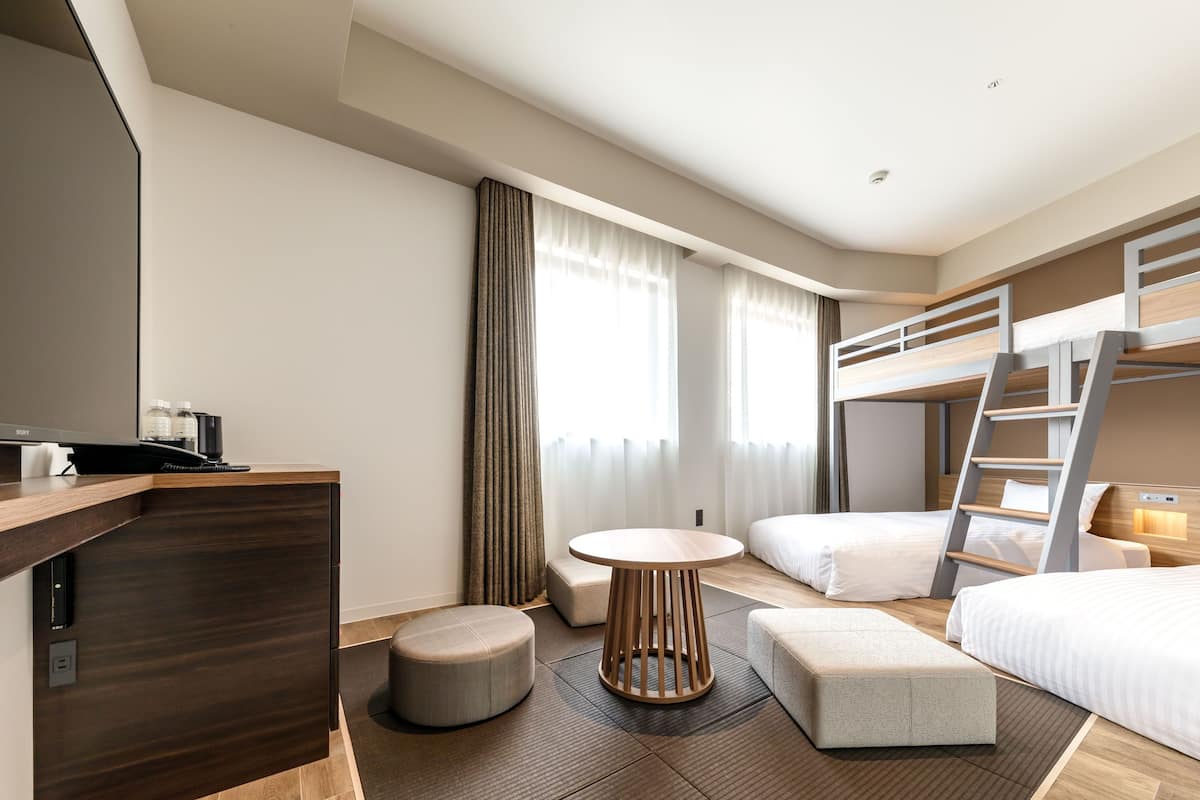 Ein modernes Hotelzimmer mit Etagenbett, Schreibtisch, Flachbildfernseher, rundem Tisch und kleinem Hocker.