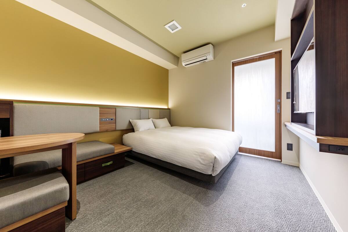Ein kompaktes Hotelzimmer mit einem Bett, einem kleinen Schreibtisch, einem Stuhl und einer eingebauten Bank.
