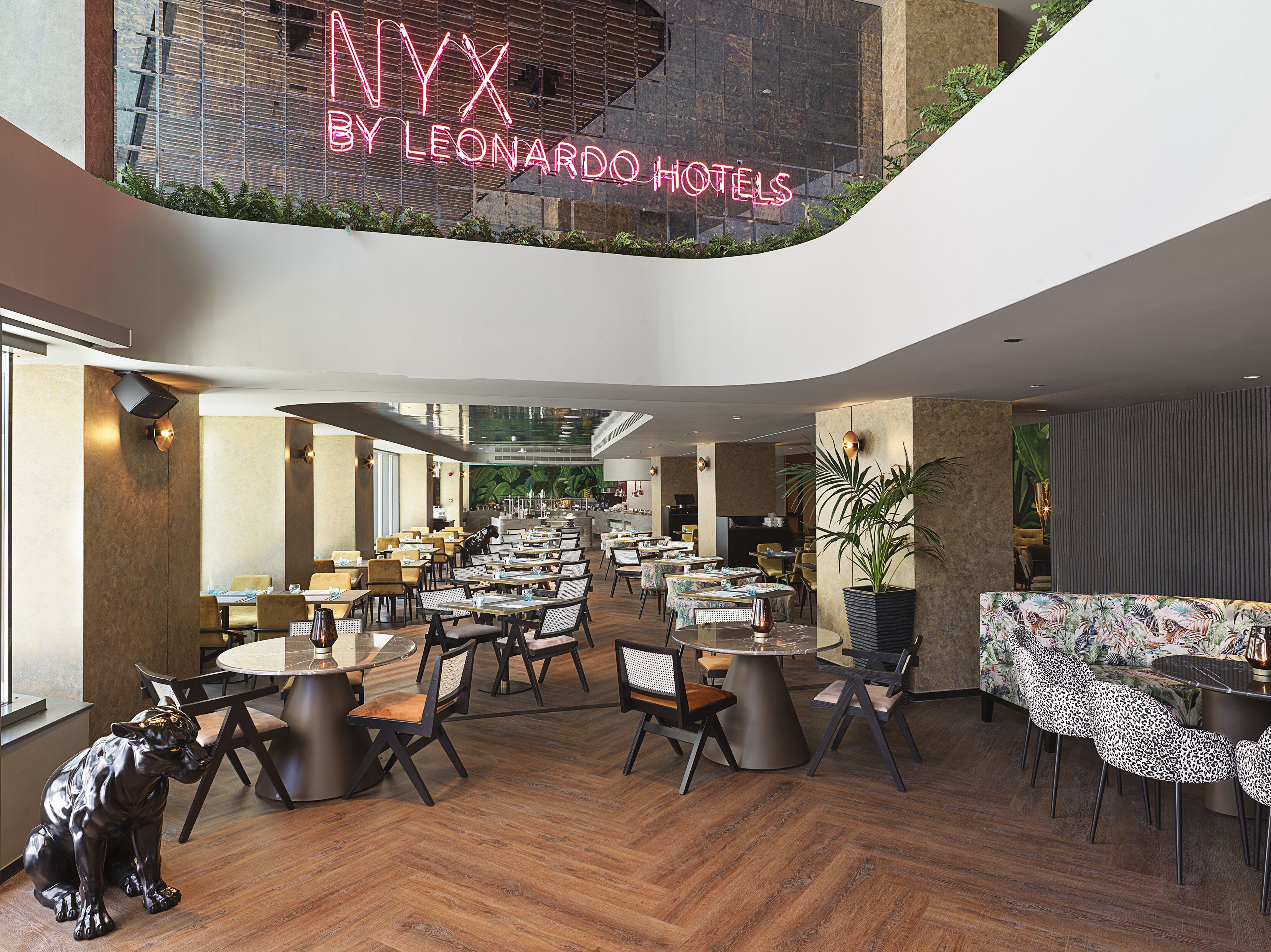 A modern hotel lobby with multiple dining areas, a visible bar, and a large plant.