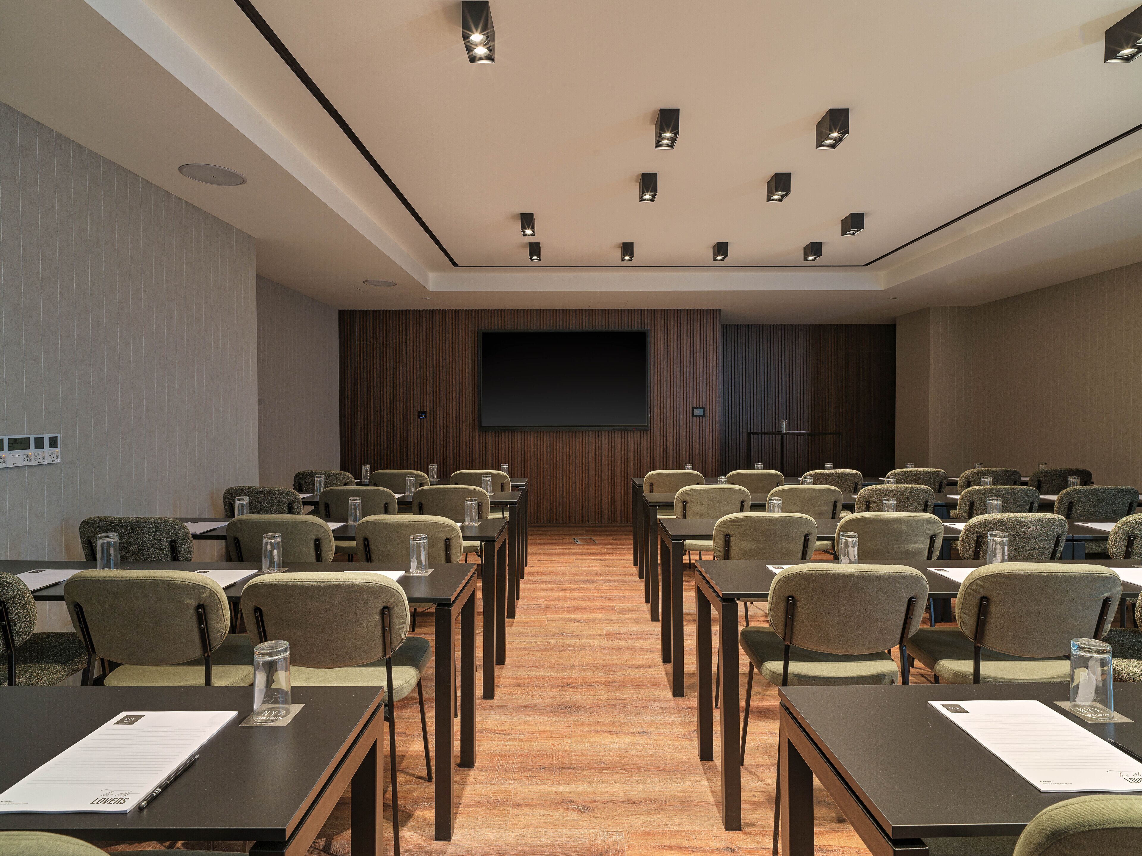 Un salón de conferencias con filas de mesas y sillas, una pantalla grande y luces en el techo.