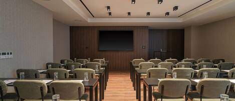 Un salón de conferencias con filas de mesas y sillas, una pantalla grande y luces en el techo.