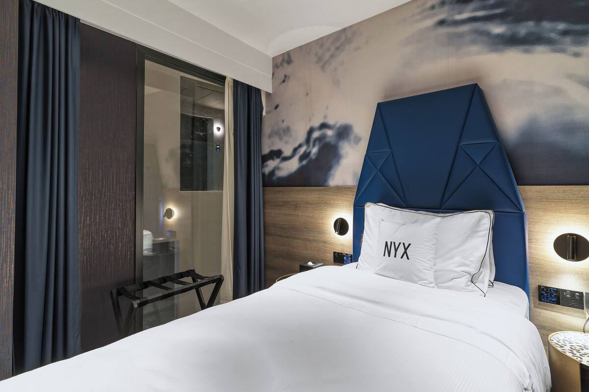 Habitación de hotel con cama, mesita de noche, pared con un cuadro y baño con puerta de cristal.