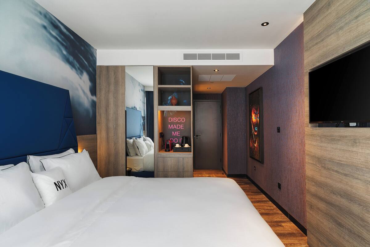Habitación de hotel moderna con una cama grande, televisión, escritorio con ordenador y baño visible a través de un separador.