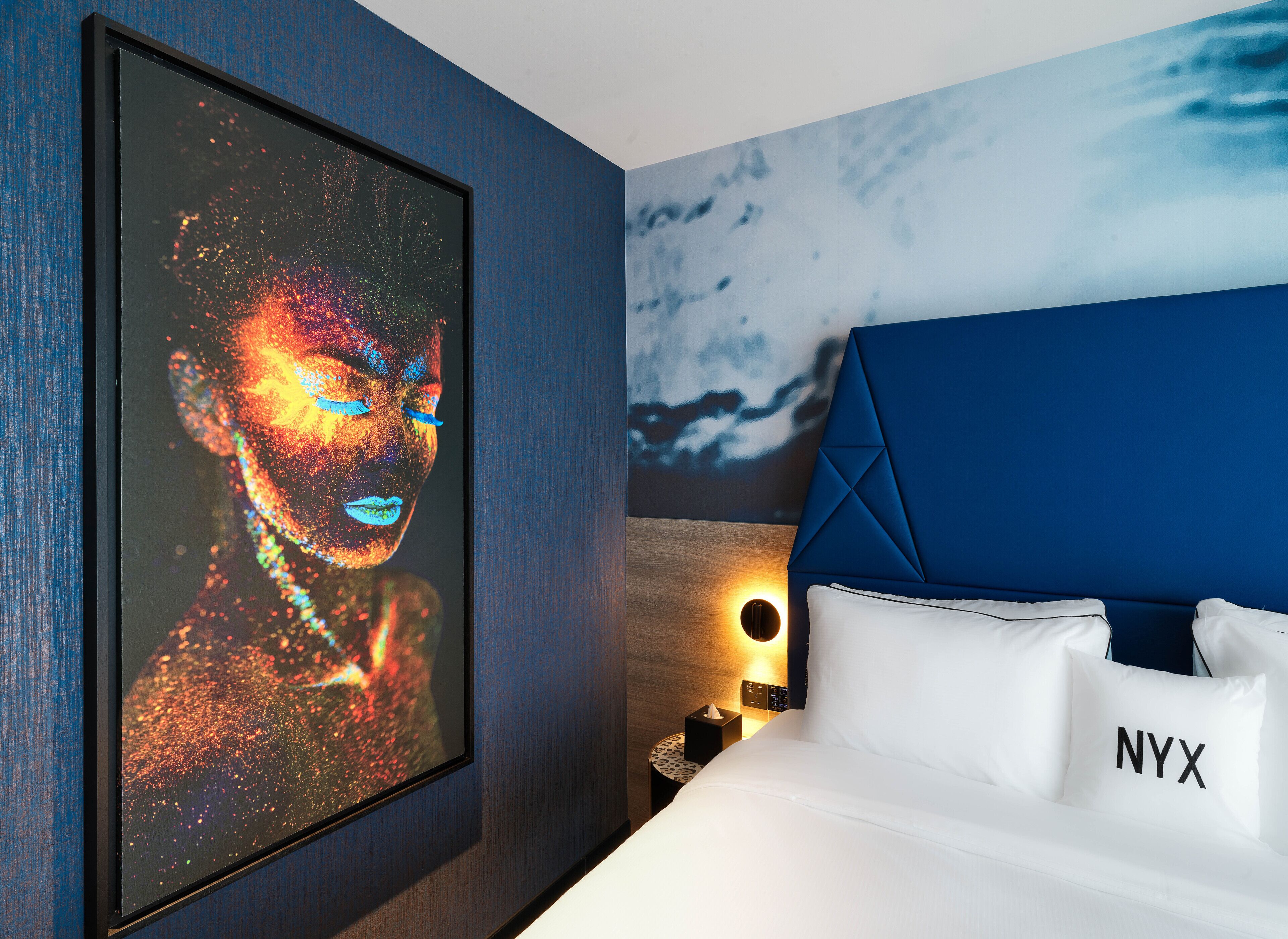 Une chambre d’hôtel moderne avec une grande œuvre d’art encadrée, une tête de lit bleue, du linge de lit blanc et une table de chevet avec une lampe.