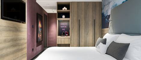 A modern hotel room with a large bed, a built-in TV, a minibar, and a wall-mounted artwork.