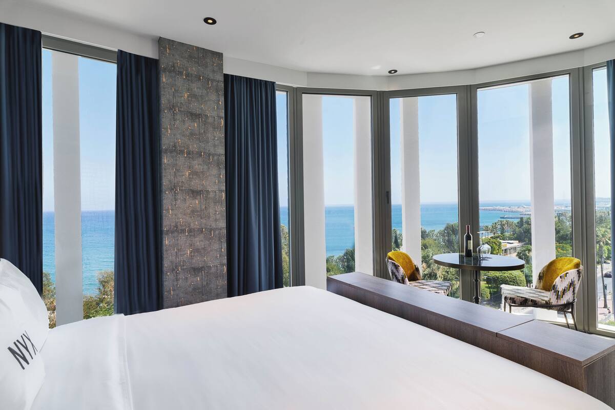 Habitación de hotel moderna con una cama grande, vistas al mar y un balcón con mesa y sillas.