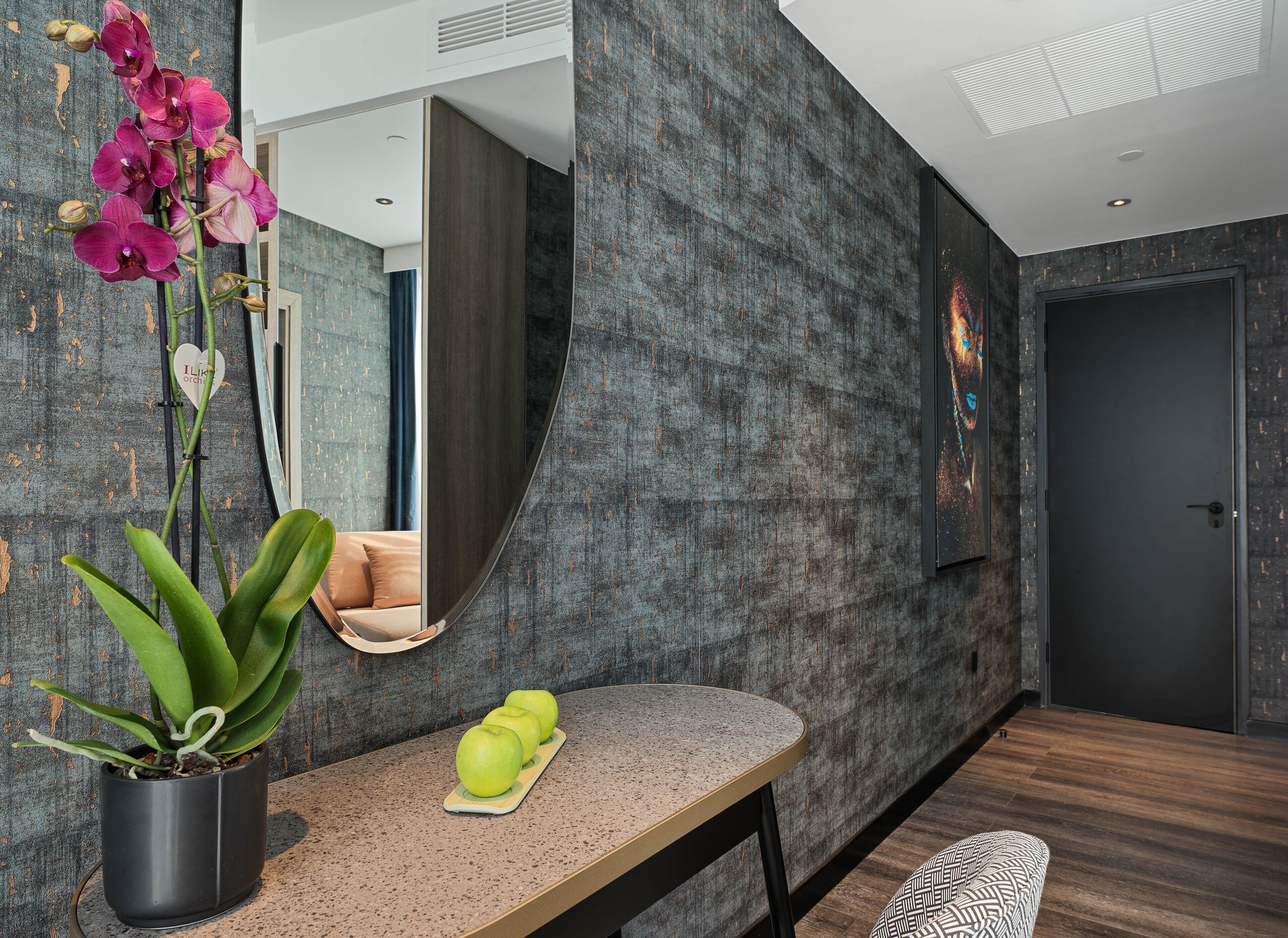 A modern interior with a dark textured wall, a round table with apples, a potted plant, and a mirror reflecting a room with a bed and a window.