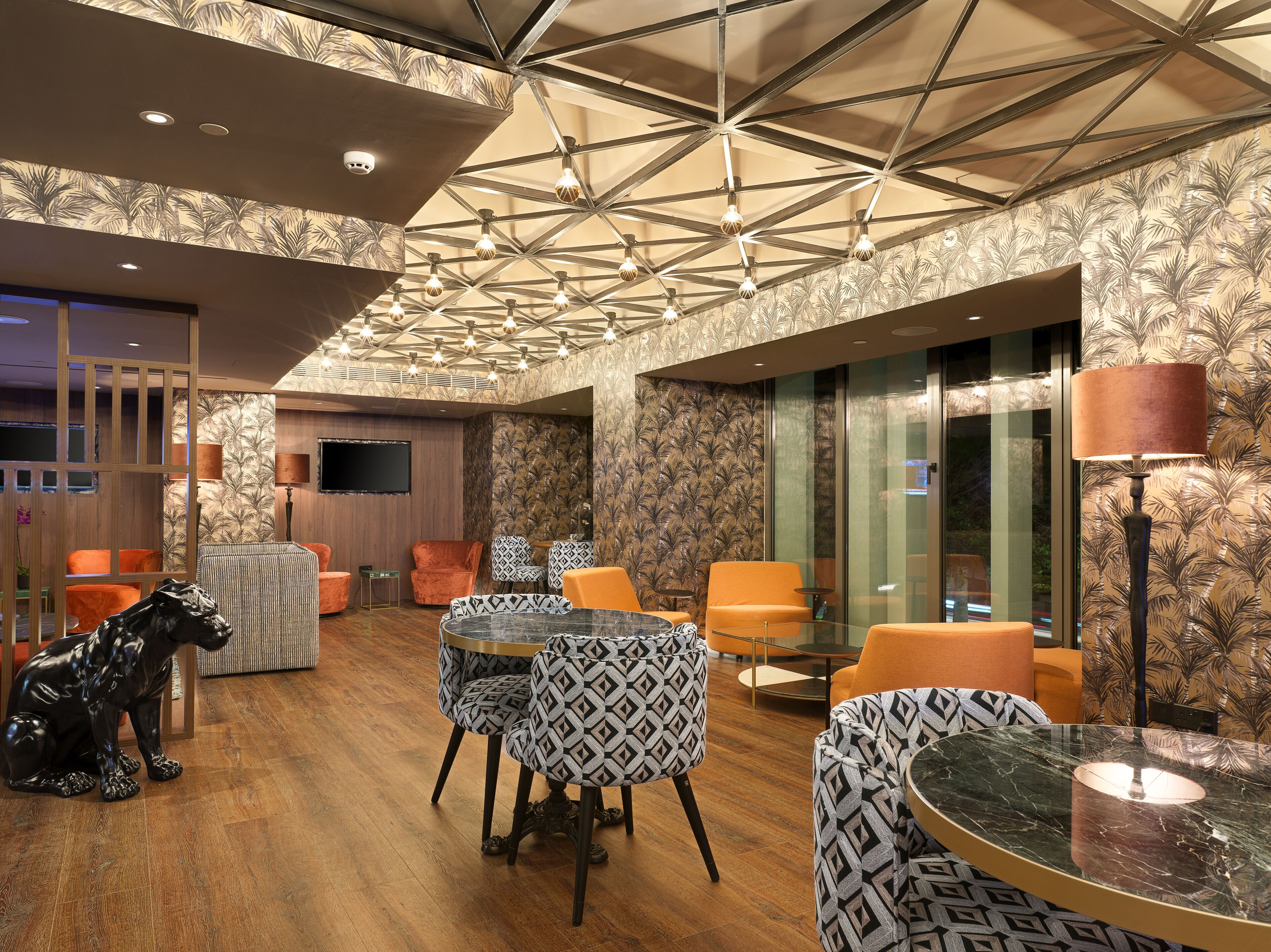 A modern lounge area with patterned wallpaper, a wooden floor, and a variety of seating options including patterned chairs and a glass table.