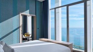 Habitación de hotel con una cama grande, vista al océano y balcón con mesa y sillas.
