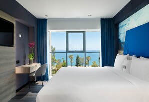 A modern hotel room with a large bed, a desk, and a view of the ocean. - NYX Hotel Limassol (Limassol)