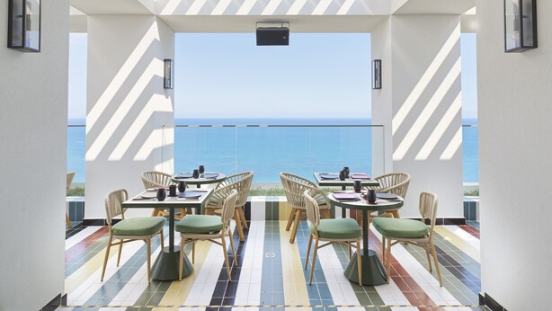 Un comedor con vistas al mar, que presenta baldosas a rayas en el techo y el suelo, sillas de mimbre con cojines, y una mesa lista para comer.