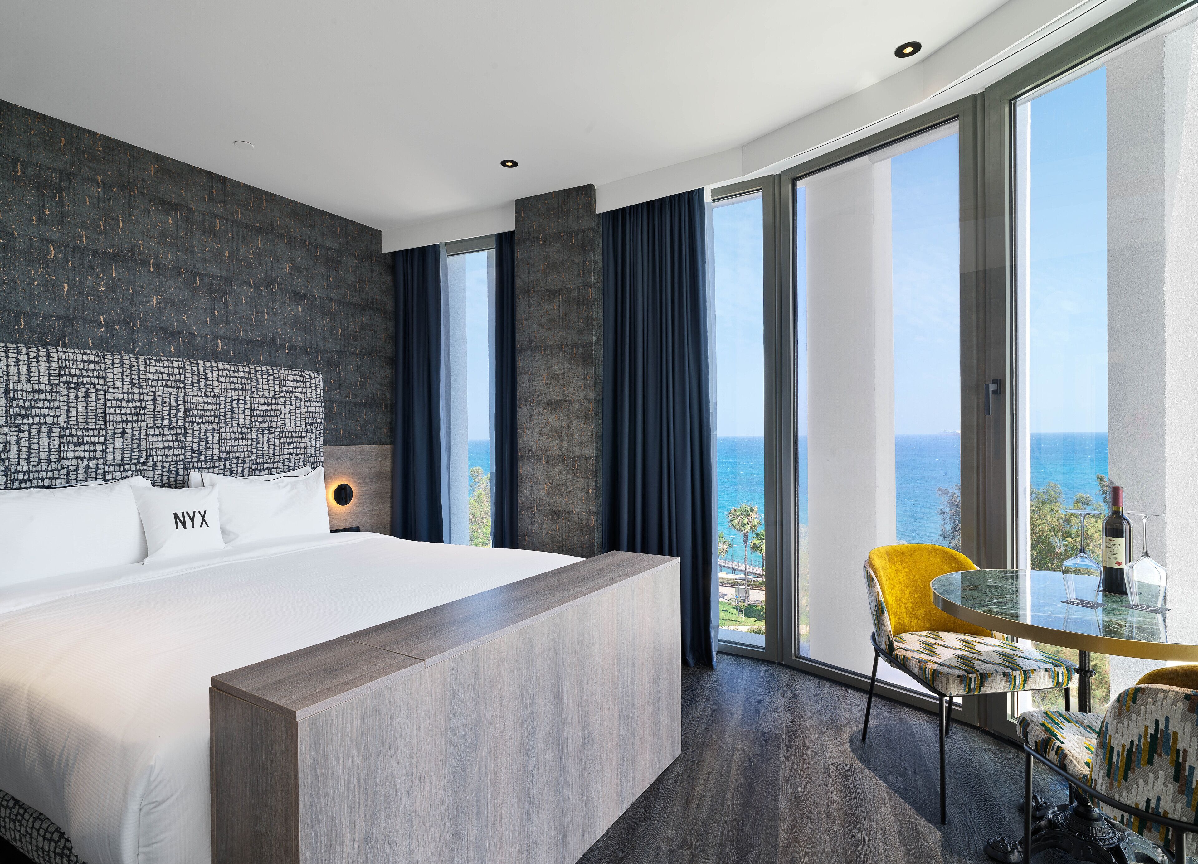 Una habitación de hotel moderna con una cama grande, vistas al mar y un balcón con mesa y sillas.