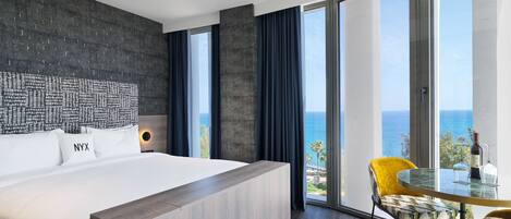 Una habitación de hotel moderna con una cama grande, vistas al mar y un balcón con mesa y sillas.
