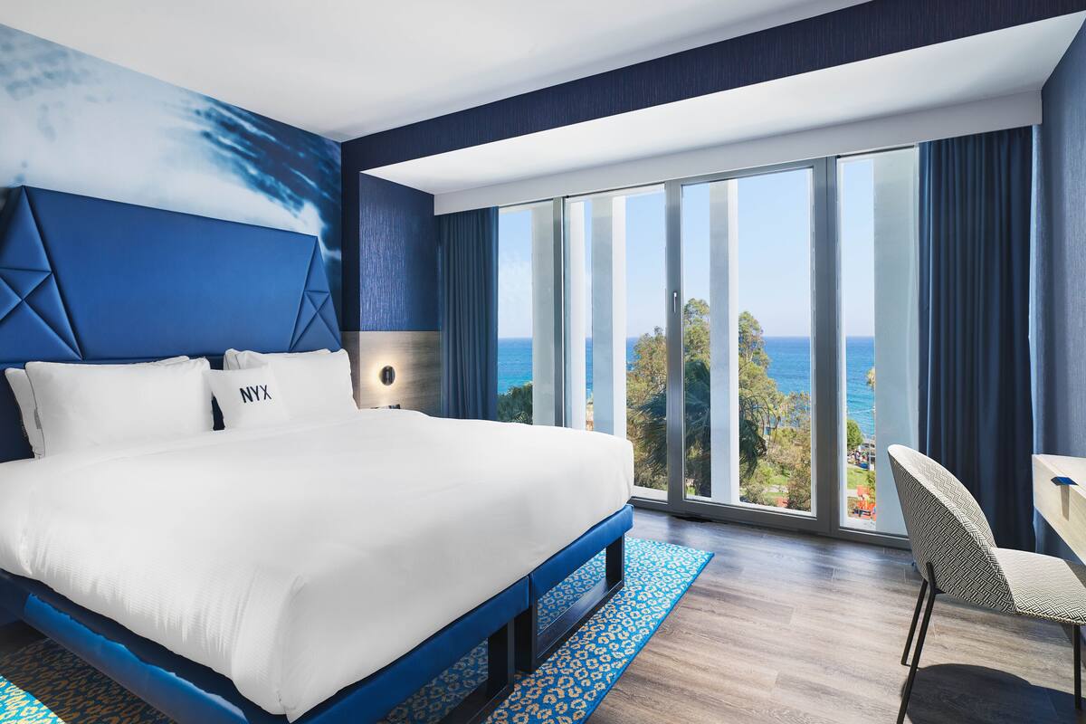 Habitación de hotel moderna con una cama grande, vista al océano y un balcón con una silla.