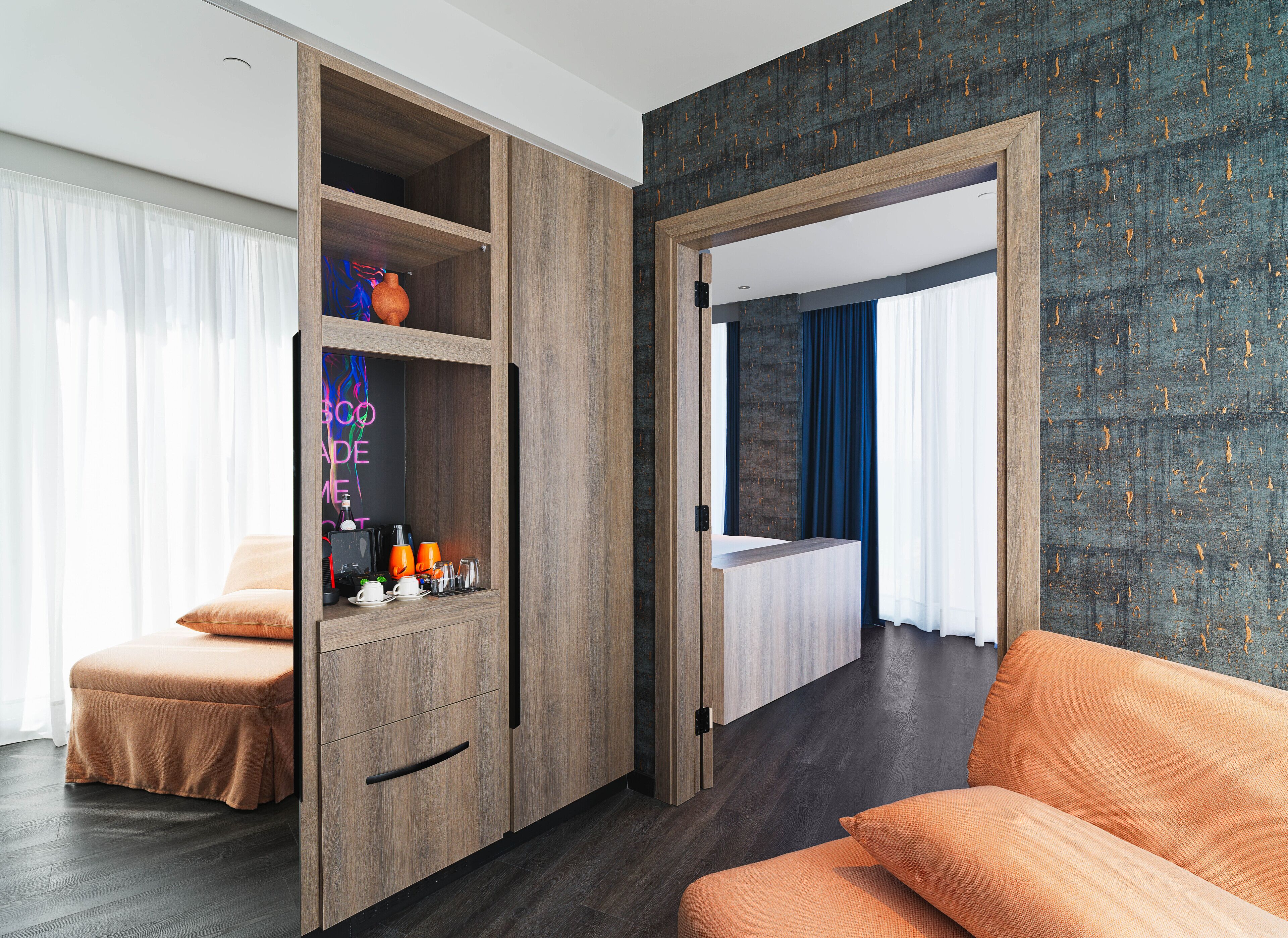 A modern hotel room with a wooden wardrobe, a bed with a dark headboard, and a view of the city through large windows.