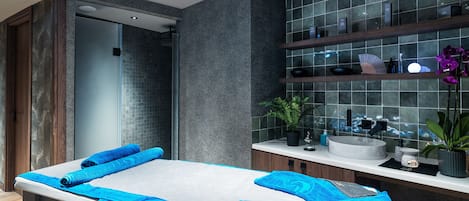 A modern spa room with a massage table, a sink, and a shower area.
