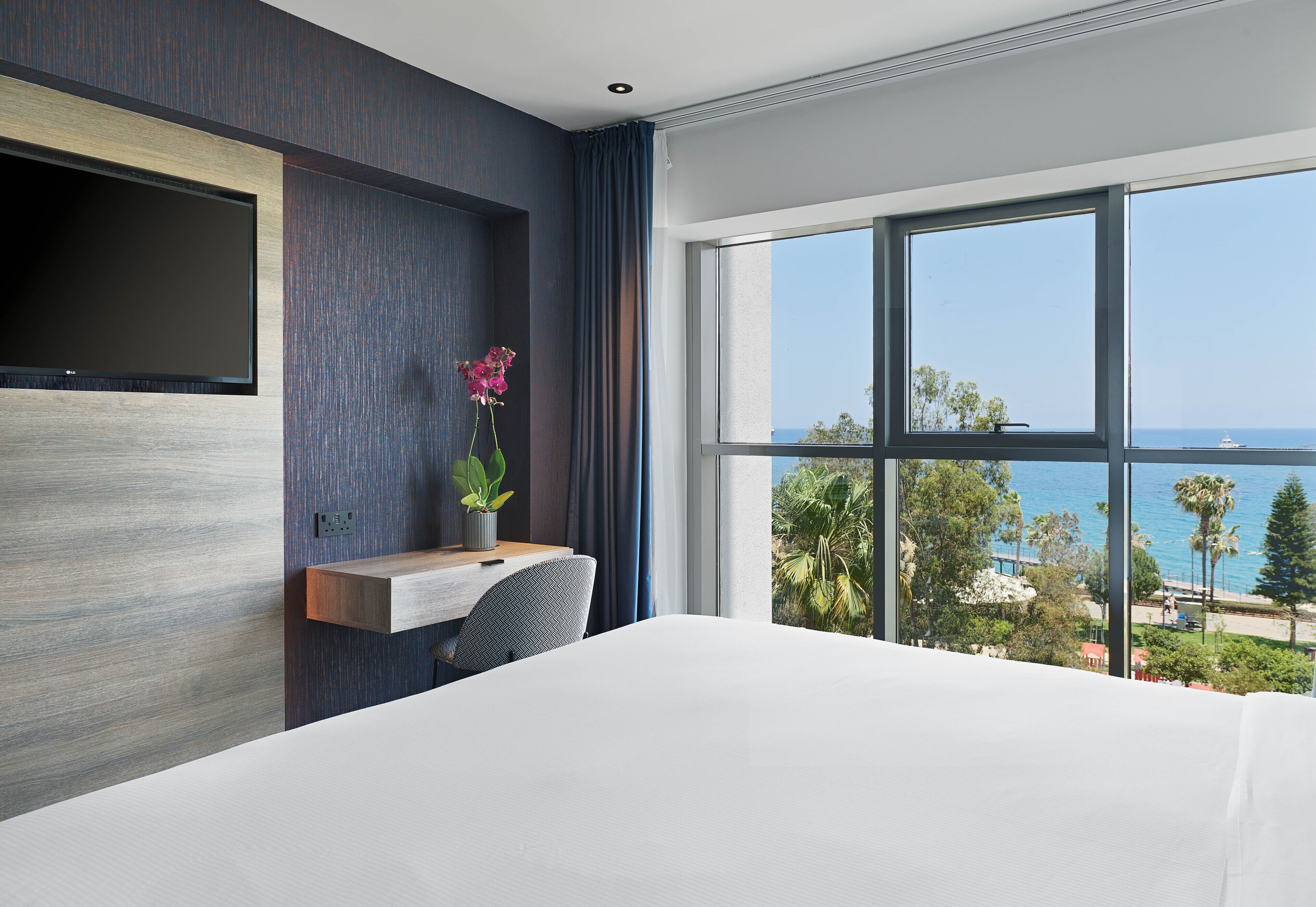 Une chambre d’hôtel moderne dotée d’un grand lit, d’un bureau avec une chaise, d’un téléviseur à écran plat fixé au mur et d’une grande fenêtre offrant une vue sur l’océan et les palmiers.