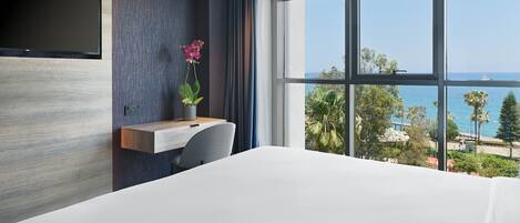 Habitación de hotel moderna con una cama grande, un escritorio con silla, un televisor de pantalla plana en la pared y un ventanal con vistas al océano y palmeras.