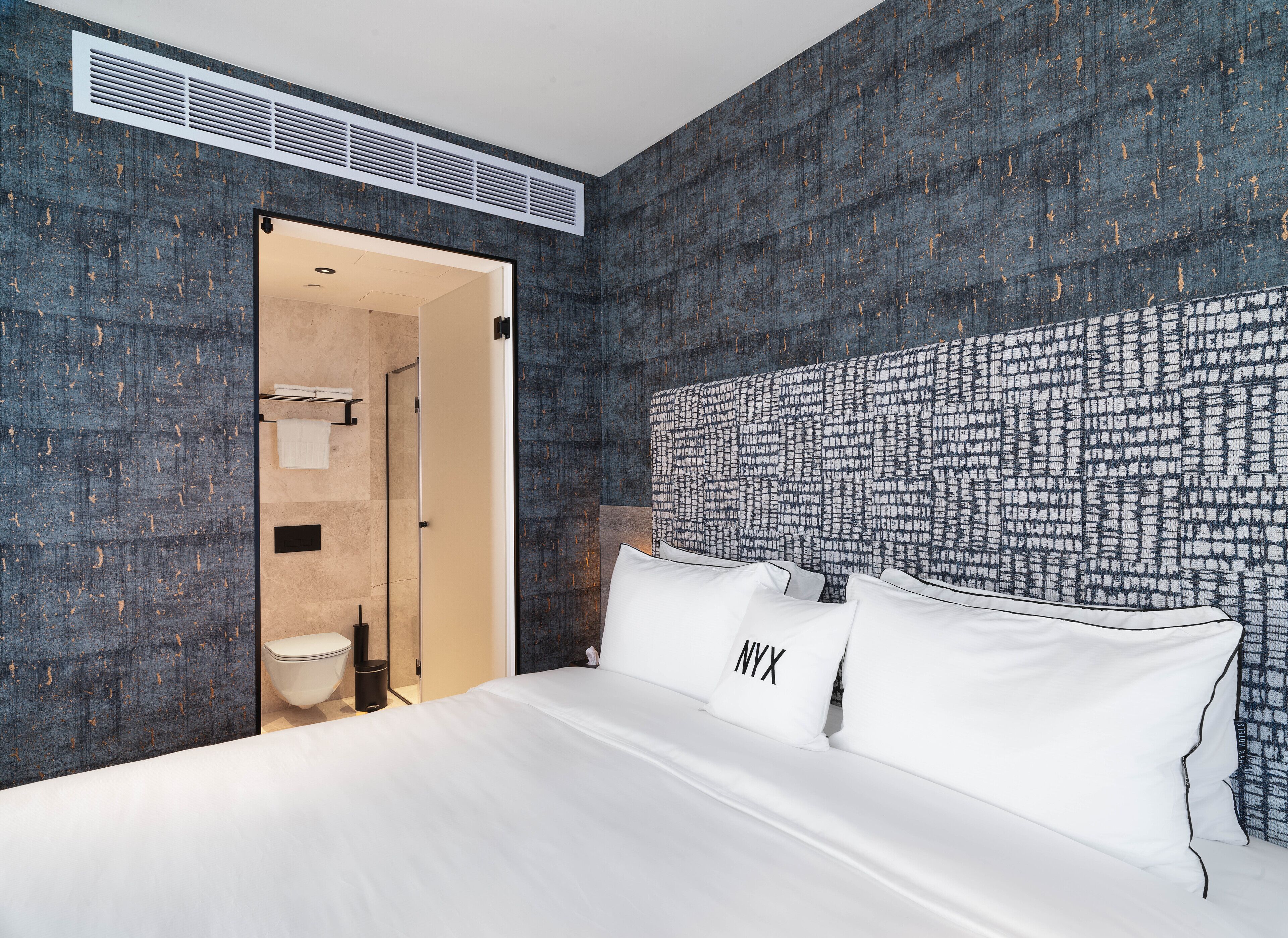 A hotel room with a large bed, a headboard with a cityscape pattern, and a bathroom visible through an open door.