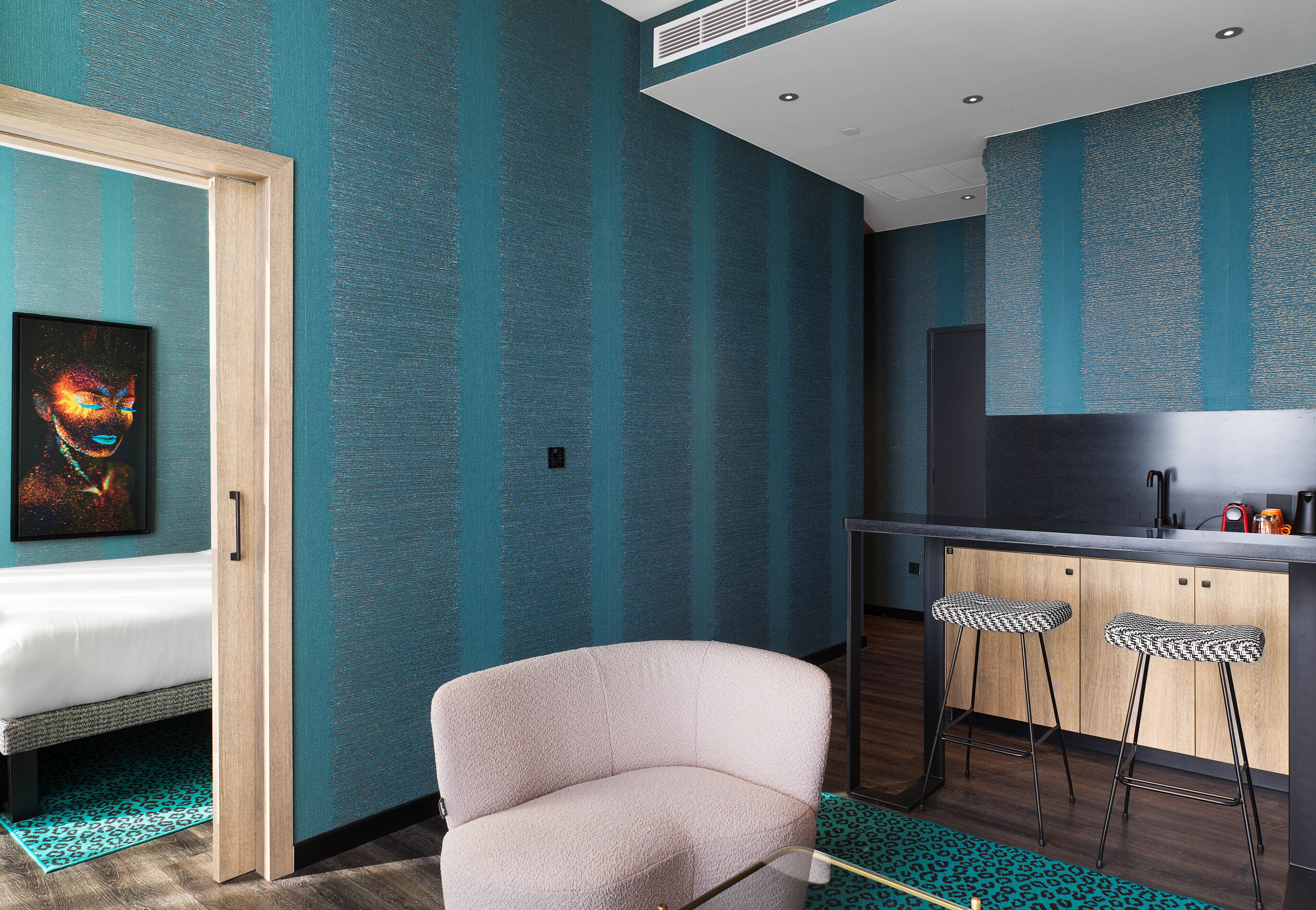 A modern room with teal striped walls, a white sofa, a kitchen area with a black countertop, and two bar stools.